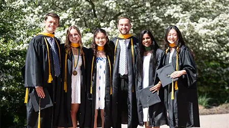 Six graduates smiling in caps and gowns outside by flowering trees.